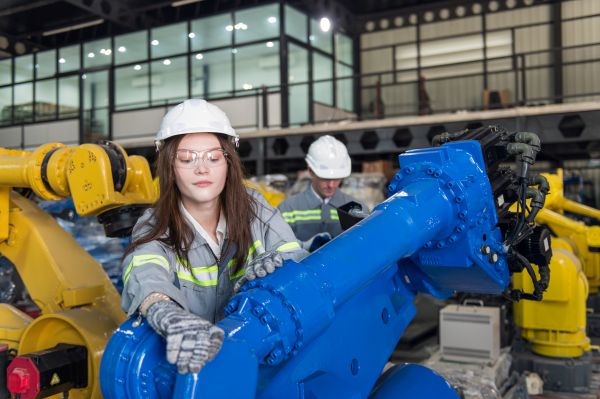 Wartung ist die Basis um effektiv Maschinenstillstand zu verhindern. Ingenieurin wartet Roboterarm während der Techniker den elektronischen Betrieb prüft